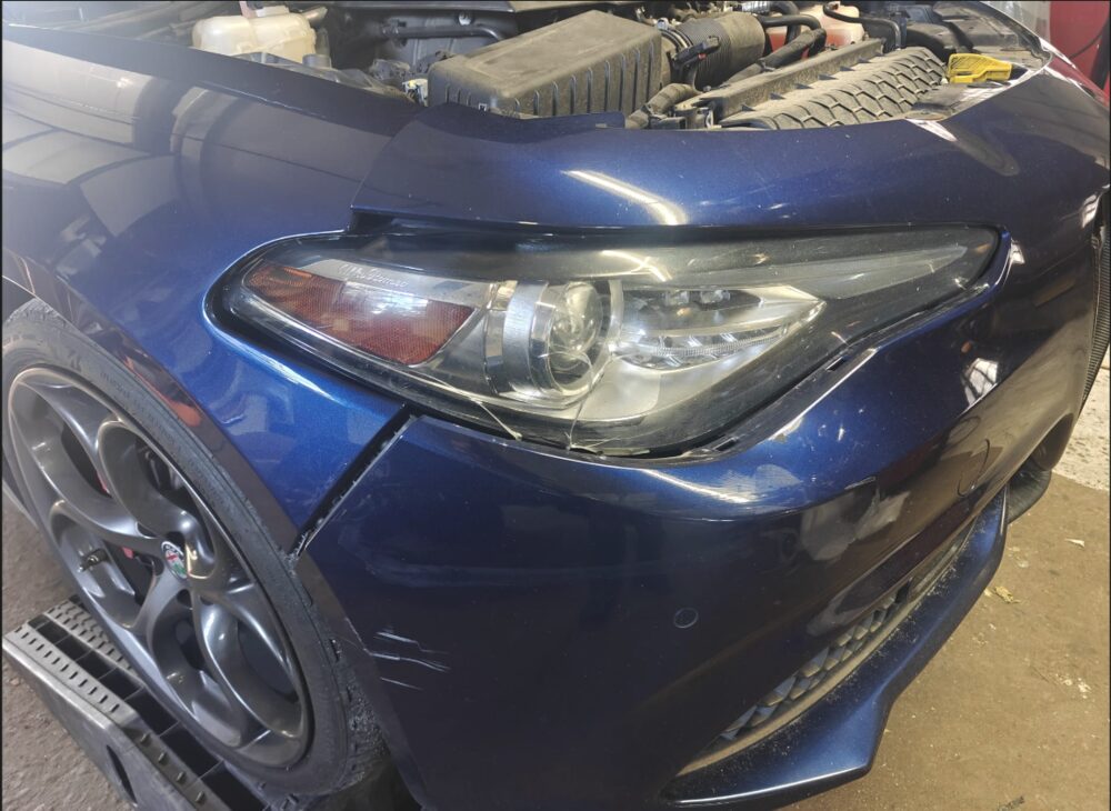 Cracked headlamp lens and damaged front bumper corner on a dark blue Alfa Romeo Giulia in the workshop at Iron Wolf Motors in Worcester, MA