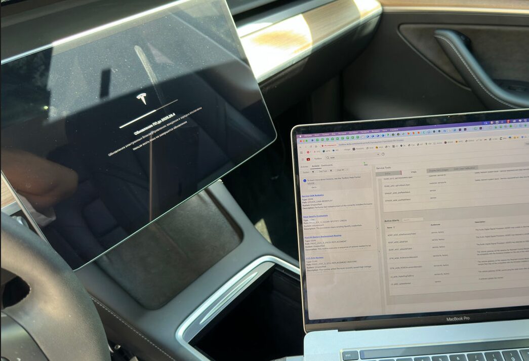 Tesla center screen running a firmware update alongside a MacBook running Tesla service software at Iron Wolf Motors in Worcester, MA