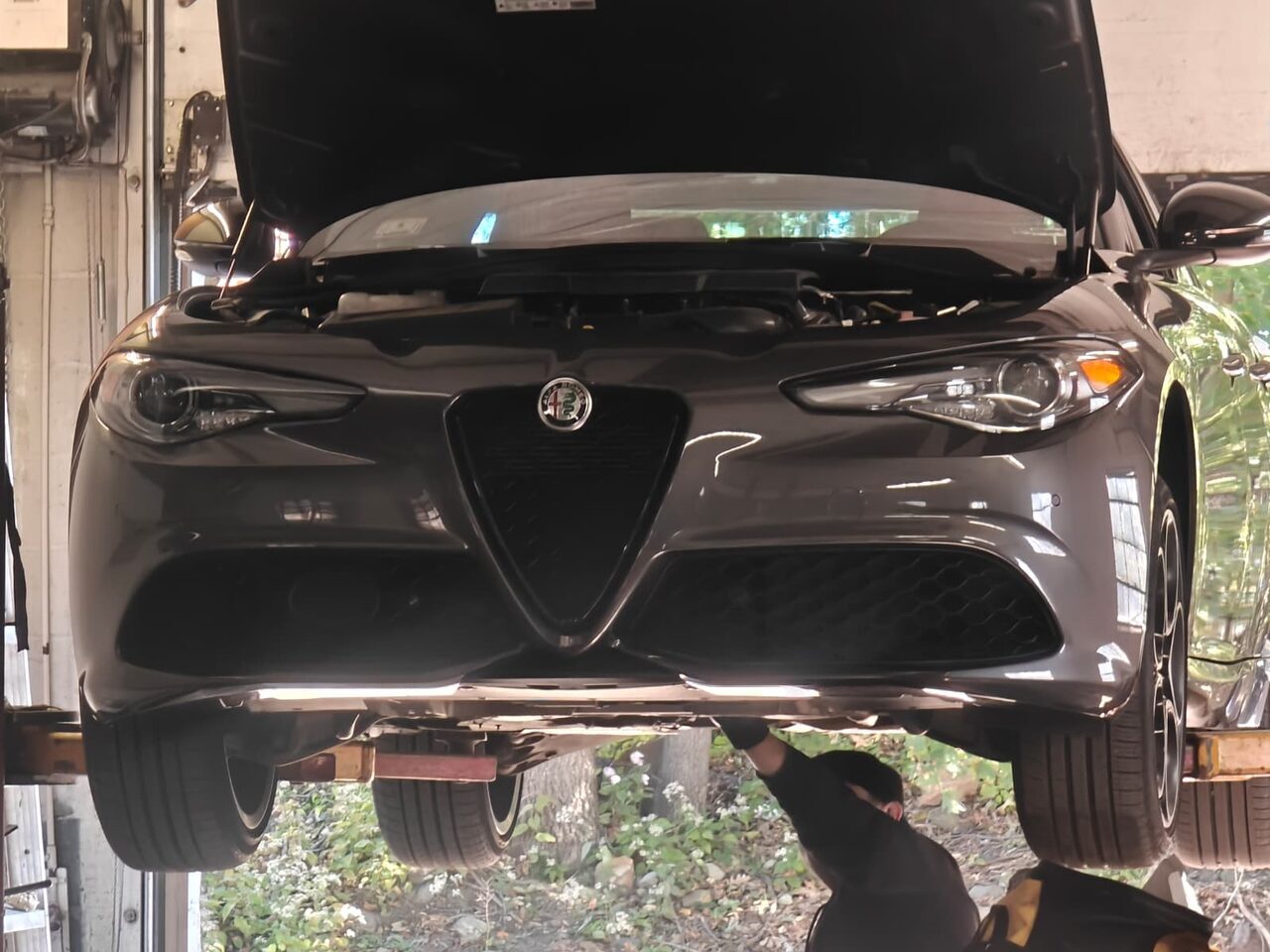 Grey 2022 Alfa Romeo Giulia raised on a two-post lift at Iron Wolf Motors in Worcester, MA, bonnet open, with a technician inspecting underneath