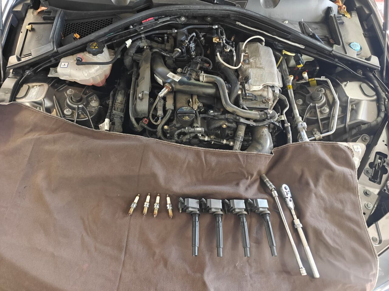 Engine bay of a 2022 Alfa Romeo Giulia 2.0 turbo with a fender cover laid over the front, four old spark plugs, four ignition coils and a ratchet with a spark plug socket lined up on the cover