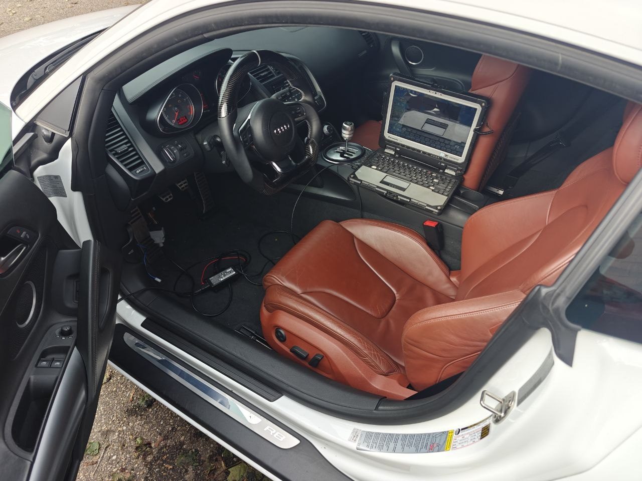 Rugged diagnostic laptop on the passenger seat of an Audi R8 with orange leather interior, cabled to the car during ECU and immobilizer programming