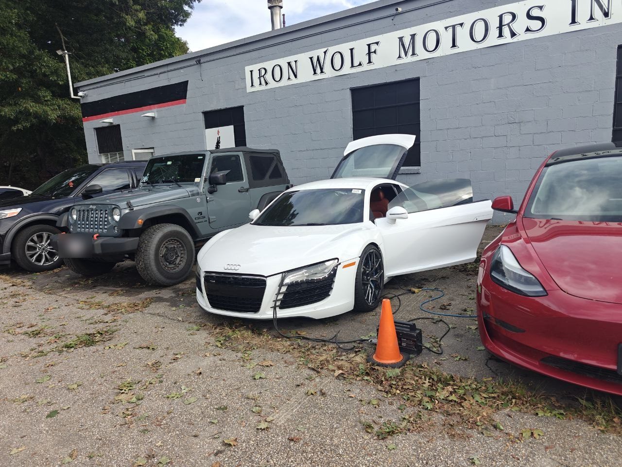 White Audi R8 parked outside Iron Wolf Motors with the driver door and engine cover open and a battery support unit on the ground connected to the car beside an orange cone