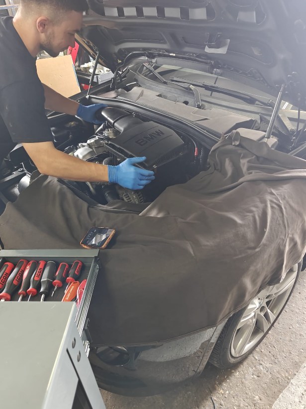 Iron Wolf Motors technician in gloves working in the engine bay of a 2015 BMW 535i, fender cover on, tools in the toolbox beside the car