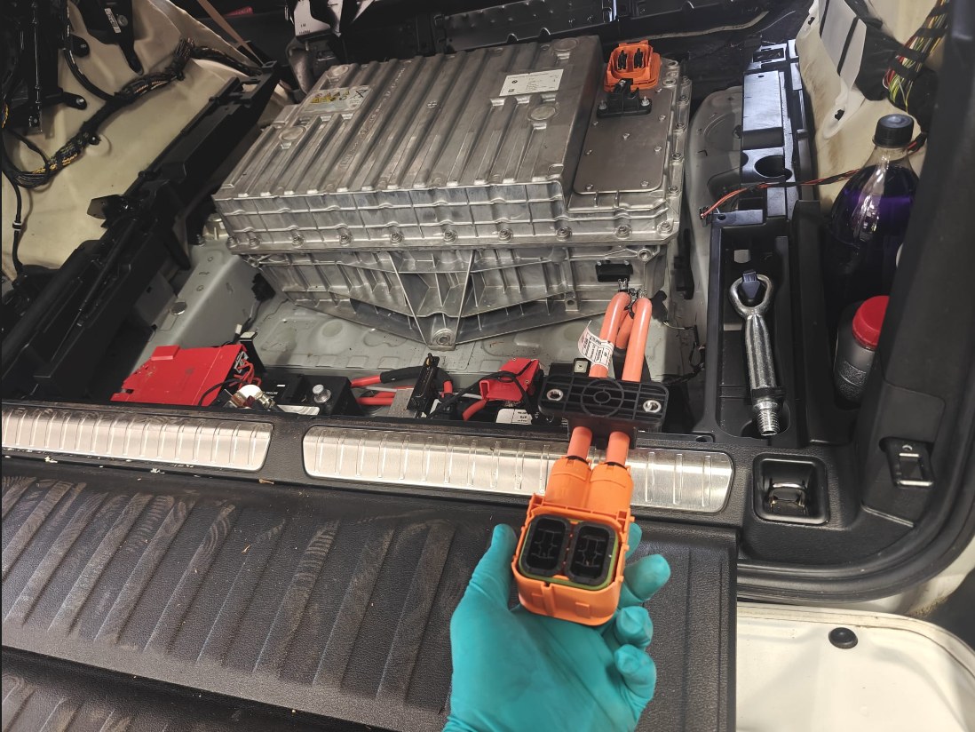 BMW X5 xDrive40e cargo area opened up with the high-voltage battery in place, a gloved hand holding the disconnected orange high-voltage connector