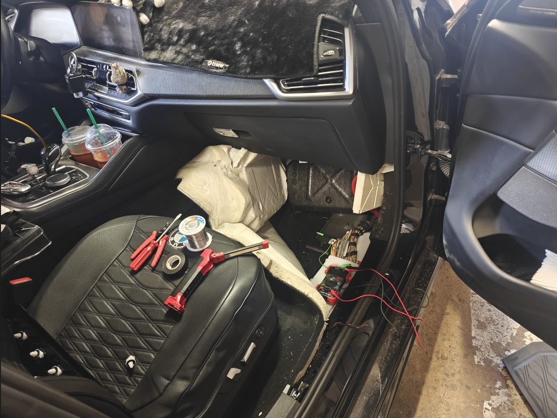Front passenger footwell of a BMW X6 with the trim removed, harness exposed, a multimeter, soldering wire and tools on the seat