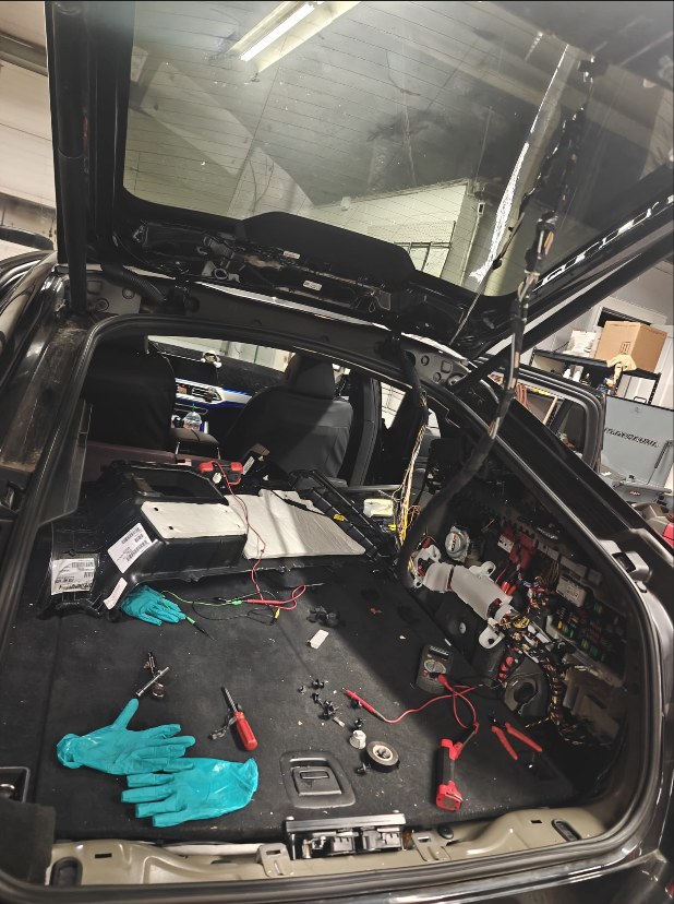 Cargo area of a 2021 BMW X6 with the trim removed, wiring and fuse box exposed, test leads and tools laid out for tracing a damaged harness
