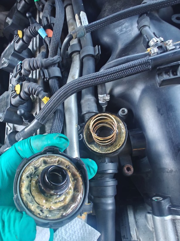 Failed PCV valve cover from a Maserati Ghibli held in a gloved hand, its inside coated in oily yellowish sludge, next to the open valve housing with its spring on the engine