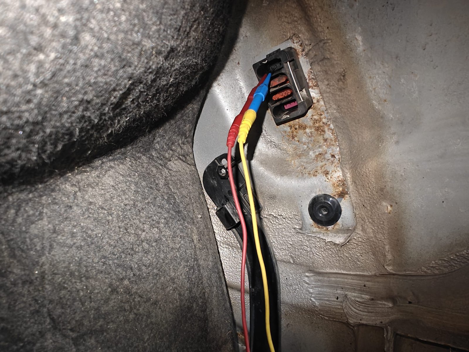 Corroded electrical connector on the rear body of a Mercedes E320 wagon, terminals with green and orange corrosion, test leads back-probing it, rust on the panel around it