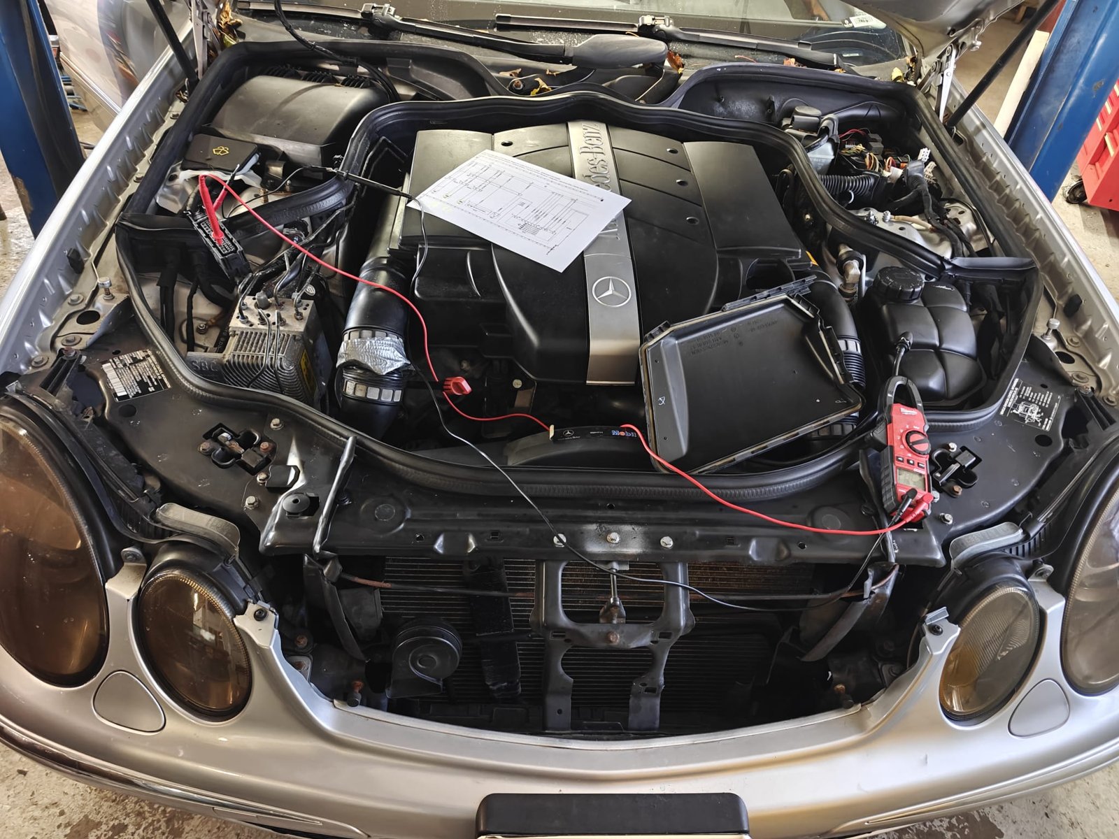 Engine bay of a silver Mercedes-Benz E320 wagon with a printed wiring diagram on the engine cover, test leads back-probing the SBC brake unit connector and a clamp multimeter on the radiator support