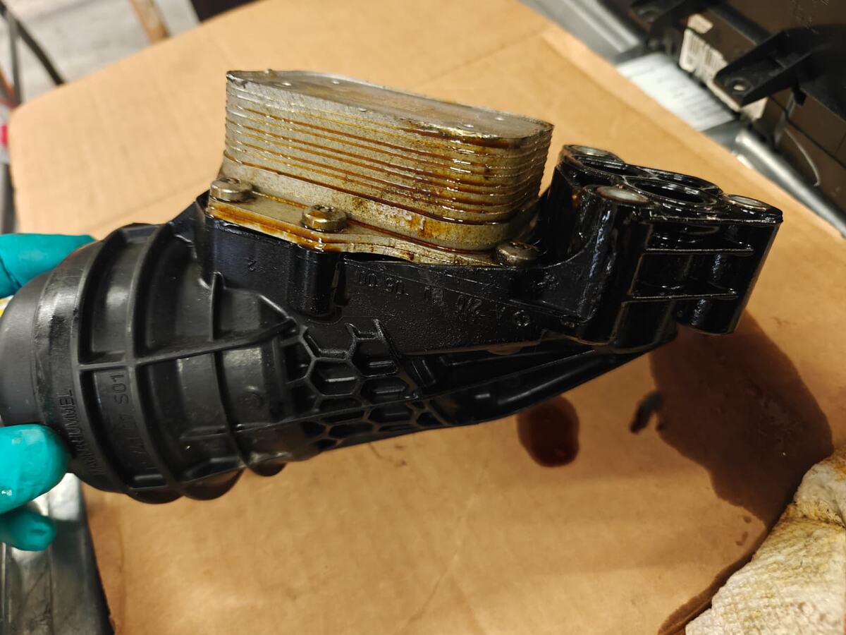 Removed oil filter housing and integrated plate-type oil cooler from a Mercedes-Benz GLA250 held over a workbench, with engine oil weeping out from between the cooler plates and running down the face of the stack