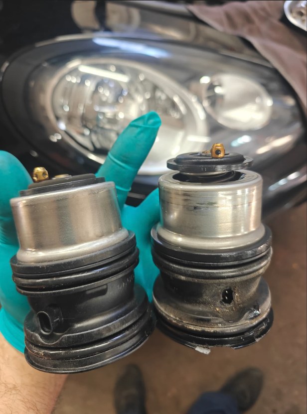 Gloved hand holding the new OEM coolant thermostat next to the old, dirty thermostat from a Porsche Panamera, in front of the car’s headlight