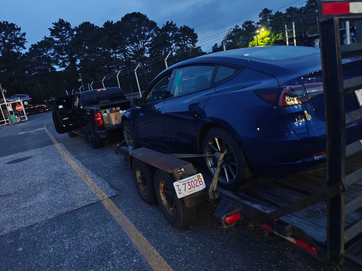 Dark blue Tesla Model 3 strapped down on a flatbed trailer at dusk, delivered to Iron Wolf Motors in Worcester, MA after a complete high-voltage shutdown