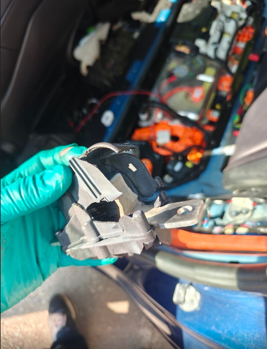 Gloved hand holding the removed pyrotechnic fuse assembly from a Tesla Model 3, with the opened high-voltage compartment and orange HV cabling visible behind it