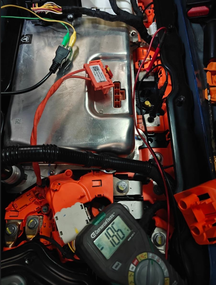 High-voltage penthouse area of a Tesla Model 3 with orange HV cabling and connectors exposed, Tesla diagnostic breakout leads attached and a multimeter probing the circuit during pyro fuse testing