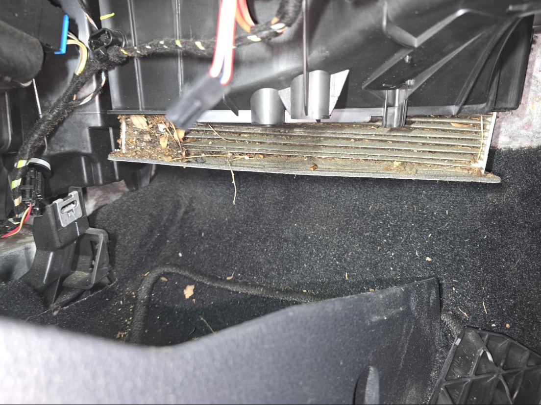 Clogged cabin air filter partly pulled out of the HVAC housing in the passenger footwell of a 2022 Volkswagen Passat, its pleats packed with leaves, seeds and dirt